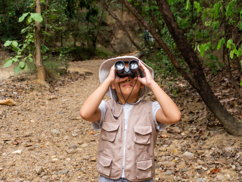 Little Asian girl wearing safari hat and vest looking through binoculars on forest nature trail
