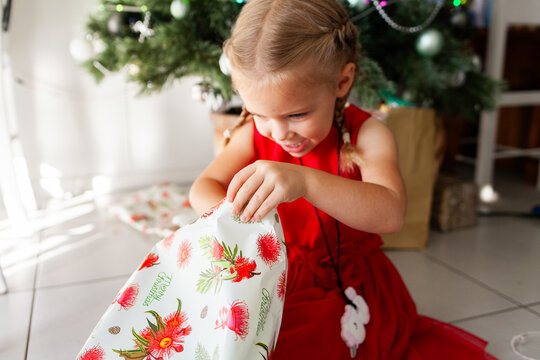 Little girl in red dress unwrapping Christmas present under tree at home