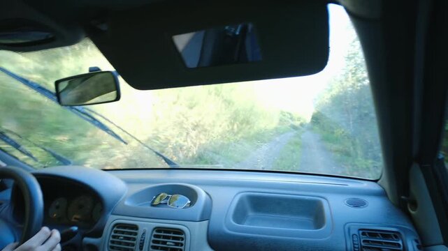 Confident young man driving car on bumpy forest road, using wipers spraying water to clean windshield. View inside vehicle, dashboard, green scenery. Young man driving car on countryside dirt road