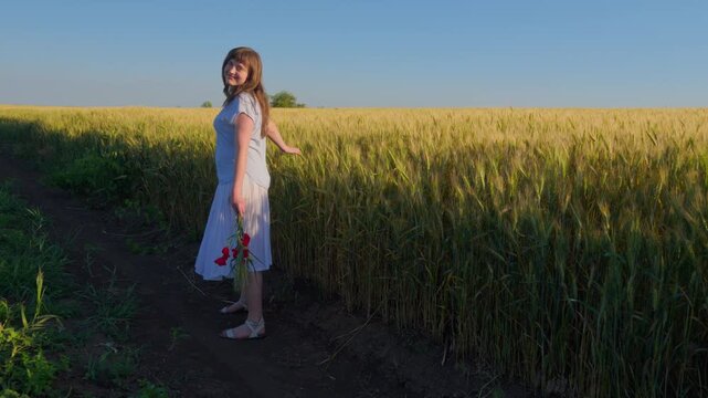 A young female teenager walking away along a path in a golden grain field while carrying a bouquet of red poppy flowers under a clear blue sky
