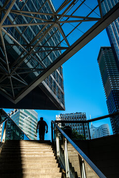 Stairway beneath a steel canopy frames a small pedestrian silhouette against blue sky and modern towers in a dramatic urban architectural scene