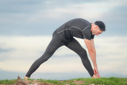 Man stretching exercise fitness flexibility warmup athlete outdoor grass sky hamstring training, focused adult male warming up on grassy field under cloudy sky before workout