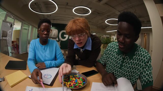 Diverse young adult coworkers analyzing candies on table during creative brainstorming session in office, captured with fisheye tilt down. Ideal for corporate training and teamwork promotions