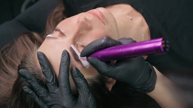 Permanent makeup artist applies white mapping paste to outline eyebrow shape before a cosmetic tattoo procedure in a professional beauty studio.