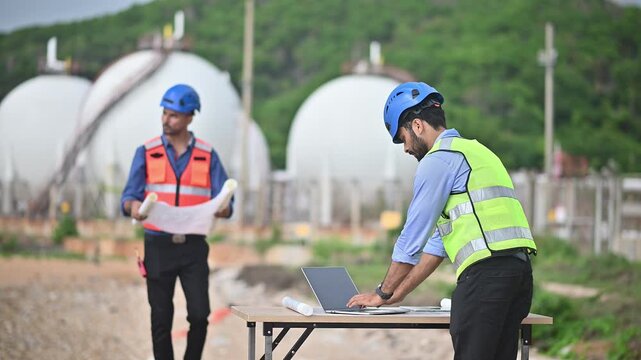 Engineers wearing safety gear, including hard hats examining survey a large blueprint tablet standing industrial facility gas or oil refinery engaged in a job requires high safety standards concept.	