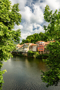 Amarante, Douro Valley, Portugal River flows calmly through charming town. Lush green trees frame scenic view. Colorful houses line riverbank with red rooftops. Sky filled with fluffy white clouds abo