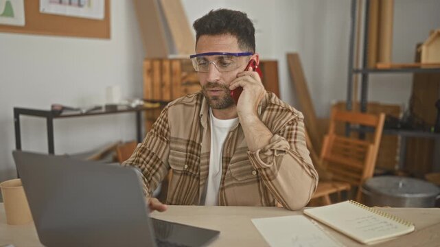 Man carpenter wearing goggles holds smartphone to ear while typing on laptop and jotting in notebook amid wood planks and tools in a studio; concentration productivity.