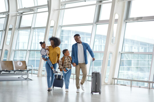 A smiling African American family with two children walks through a modern airport terminal, pulling suitcases and carrying bags