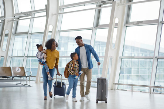 A Black family with two young children walks through a modern airport terminal, pulling suitcases and carrying bags, ready for their journey