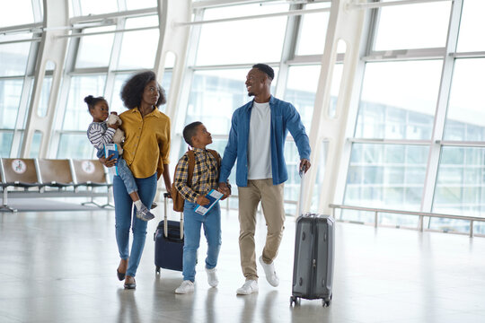 A smiling Black family with two children walks through a modern airport terminal, holding passports and pulling suitcases for their vacation