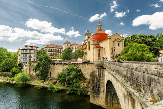 Amarante, Douro Valley, Portugal Old stone bridge spans calm river under blue sky. Historic church with red roof stands on hillside. Trees line riverbank, adding green contrast. Buildings with warm-to