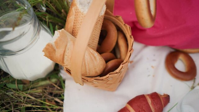 Close up of woven basket filled with bread rolls and bagels placed on white fabric next to glass milk bottle. Hand reaches into frame selecting item from picnic spread set against grassy