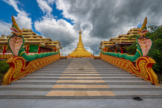 Golden Pyay Tha Lin Aye Pagoda, known as Phra That Tagaw, features a wide staircase with naga handrails leading toward the main stupa under a blue cloudy sky in Myanmar.