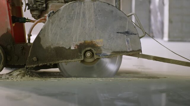 Concrete saw makes expansion joints on commercial slab floor. Slurry splashes around spinning circular cutting blade during floor work