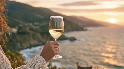 Naklejka premium Woman Hand Holding White Wine Glass Over Scenic Ocean Coast Sunset View Summer Evening Landscape.