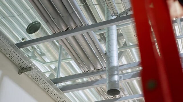 Vertical air duct drops down from busy ceiling utility setup. Overhead infrastructure features insulated piping and sturdy steel framing