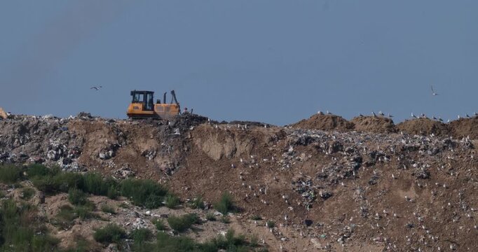 Bulldozer and unrecognizable operator on MSW landfill with a dump truck backing up to unload with atmospheric heat shimmer slow motion