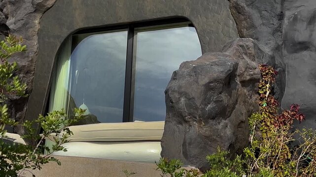 Scenic view of arched window set in volcanic rock in Santorini Greece. Green foliage surrounds the stone structure while the glass reflects the cloudy coastal landscape.