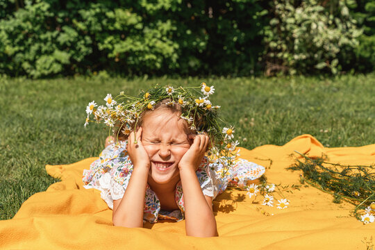 Happy little girl squints in sun and lies on yellow picnic blanket, wearing wreath of wildflowers, on bright, sunny summer day. Joyful childhood, playing outdoors, carefree summer fun in nature