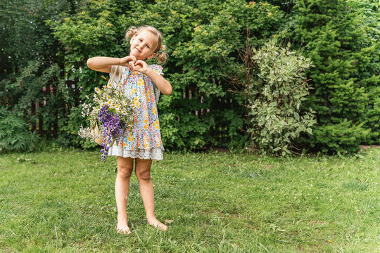 Little girl in floral dress and barefoot on green lawn, holding bouquet of wildflowers and making heart gesture with her hands, against backdrop of dense green trees. Children's happiness love