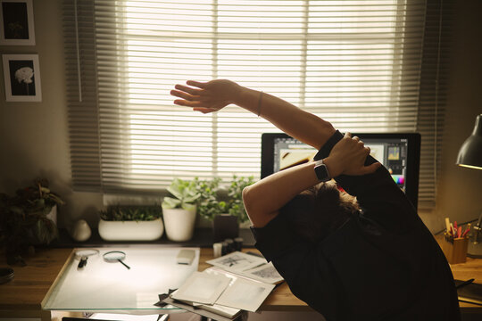 Rear view of young man stretching arms at desk in home office, taking break from computer work. Concept of remote work wellness, suitable for healthy lifestyle marketing