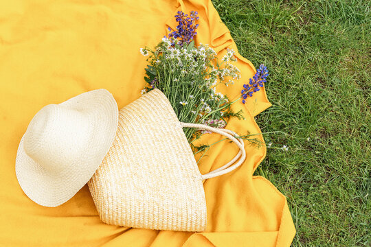Woven straw bag and straw hat lie on bright yellow blanket on green grass, next to bouquet of wildflowers (daisies, lupines, and wild herbs), viewed from above