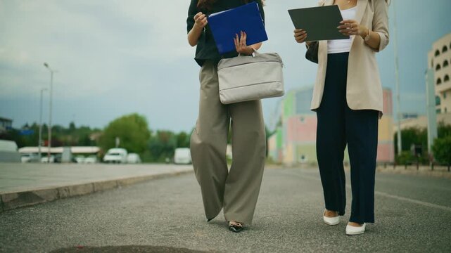 Two professional businesswomen walk purposefully together outdoors in an urban setting. These corporate colleagues commute or head to a meeting on a city street.