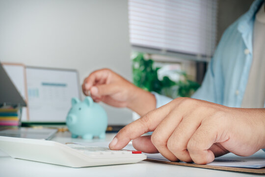 Person calculating finances with calculator and dropping coin into blue piggy bank on office desk, concept of financial planning, saving money, budget management, and investment success.