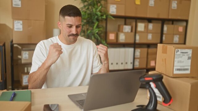 Man at laptop raises fists in building packing office amid boxes and scanner; small business success joy.