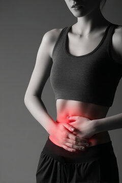 A woman stands in a studio with a gray background. She wears a gray top and black pants and holds her stomach with both hands, showing discomfort