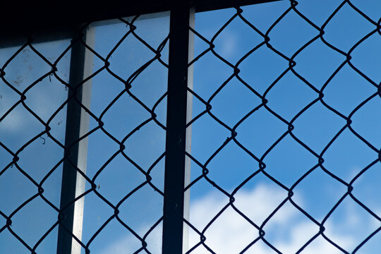 Metal chain link wire mesh fence against a bright blue sky with soft white clouds symbolizing freedom