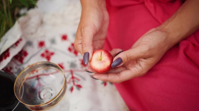 Close up of woman hands holding and examining small red fruit with purple nail polish on fabric. Background includes patterned textile empty glassware grapes greenery soft lighting calm