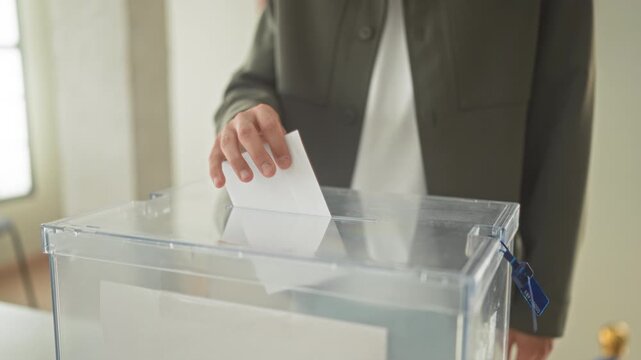Man places ballot with hand into transparent ballot box labeled vote at polling table inside building; duty civic participation.