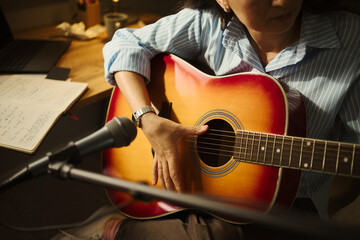 Fototapeta premium Young woman playing acoustic guitar in front of microphone, composing song at home studio. Image ideal for online music lessons, creative hobbies, and digital audio workstation ads