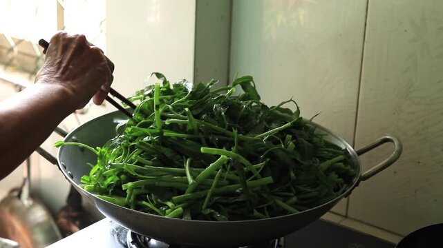 Stir frying fresh green morning glory water spinach in a hot wok using wooden chopsticks in home kitchen