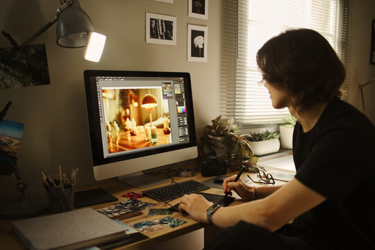 Young Asian man editing photo on computer using graphics tablet at home office. Image represents remote work and creative profession, suitable for freelance career marketing
