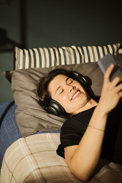 Smiling Asian young man lying in bed wearing headphones and holding smartphone. Image represents digital entertainment and youth lifestyle, suitable for mobile app marketing, vertical