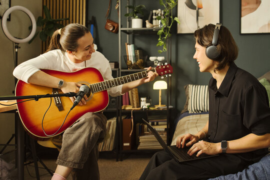 Caucasian young woman playing acoustic guitar while Caucasian young man with headphones uses laptop. Creative duo recording music podcast, suitable for home studio marketing