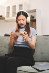 Beautiful young asian woman sitting on sofa and holding joystick playing online video game in living room at house © ARMMY PICCA
