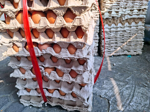 A stack of brown chicken eggs stored in grey recycled paper pulp trays, tightly bound with red plastic strapping string outdoors.