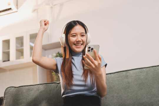 Beautiful young asian woman sitting on sofa and holding joystick playing online video game in living room at house
