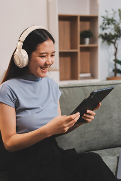Freelance Asian woman holding mobile phone typing on laptop and working from home. Happy young girl sitting on sofa in living room.