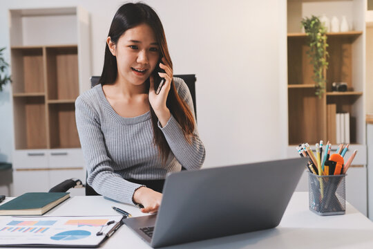 Young asian businesswoman is smiling and working on a laptop while sitting at her desk in a modern office