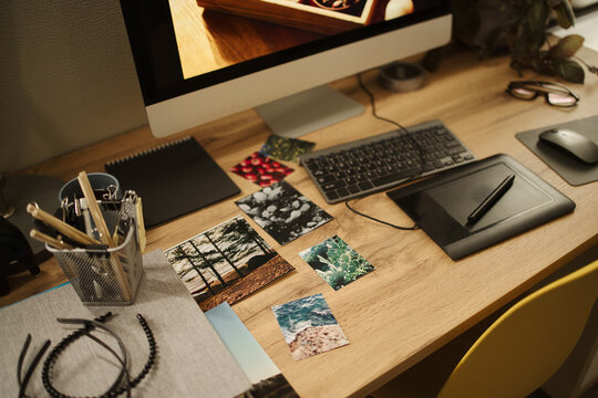 Graphic designer workspace featuring computer monitor, keyboard, graphics tablet, and printed reference photos on wooden desk, representing creative workflow, high angle shot