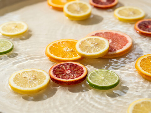 A highly realistic overhead lifestyle photograph of freshly sliced citrus fruits floating on the surface of clear, shallow water under bright natural sunlight