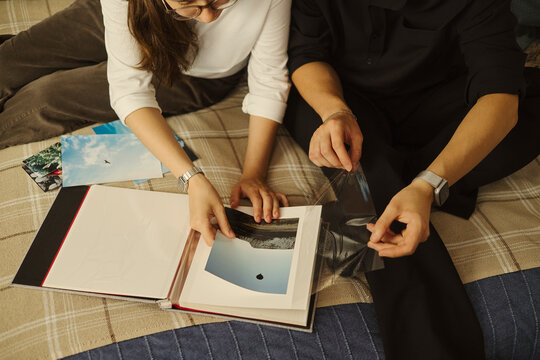 Young Caucasian woman and Asian man sitting on bed, organizing printed photos into album. Image represents memory preservation, suitable for family archiving services