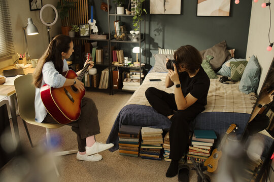 Young Caucasian woman playing acoustic guitar while young Asian man takes photos with DSLR camera in bedroom, representing content creation and hobby marketing, high angle shot