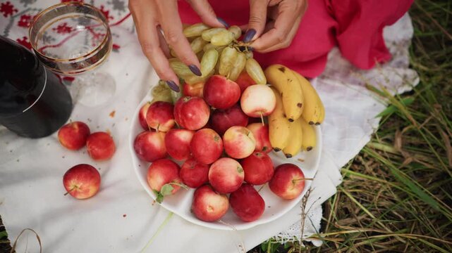 Close up of person hands arranging green grapes on top of red apples and yellow bananas in white bowl placed outdoors on grass with scattered fruit wine glass bottle pink fabric patterned
