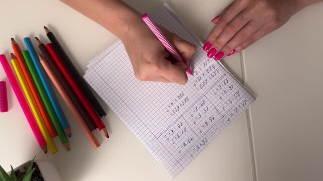 Top-down shot of hands writing math equations on squared notebook with colored pencils and pink manicure, with writing, hand, notebook, math detail, showing equation, pen, pencil, colored context