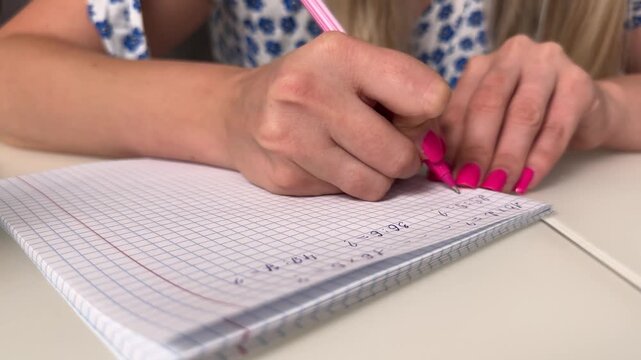 Close-up of hands writing math on squared notebook with pink pen and magenta nails, low tabletop viewpoint, with writing, hand, notebook, pen detail, showing math, equation, note, study context